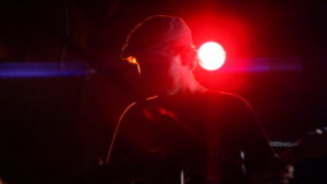A silhouette of musician in front of a bright red light, while performing on stage with a strapped musical instrument. The man is wearing a cap and has a dark shirt.
