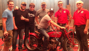 Seven smiling men from young to older, one holding a video camera, one sitting on an Indian motorcycle and all posing for the camera. Five of the younger men are wearing t-shirts. The two older men are wearing red company short-sleeved shirts.